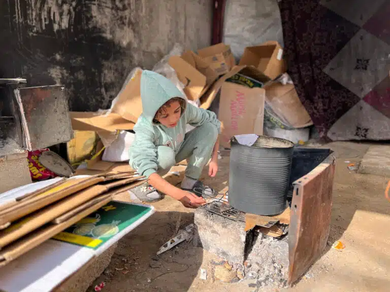 Farah, de 11 años, alimenta el fuego de una cocina improvisada tras desplazarse con su familia en la Franja de Gaza en numerosas ocasiones en los últimos dos años. Plan International y sus socios locales les han ayudado con ropa, comida y fisioterapia para su hermano © Juzoor for Health and Social Development / Plan International